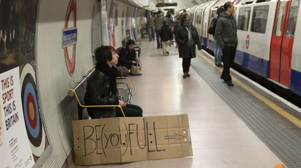 Person seated on a London Underground platform holding a BE(YOU)FULL sign, reflecting identity, awareness, and social context