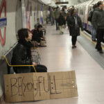Person seated on a London Underground platform holding a BE(YOU)FULL sign, reflecting identity, awareness, and social context
